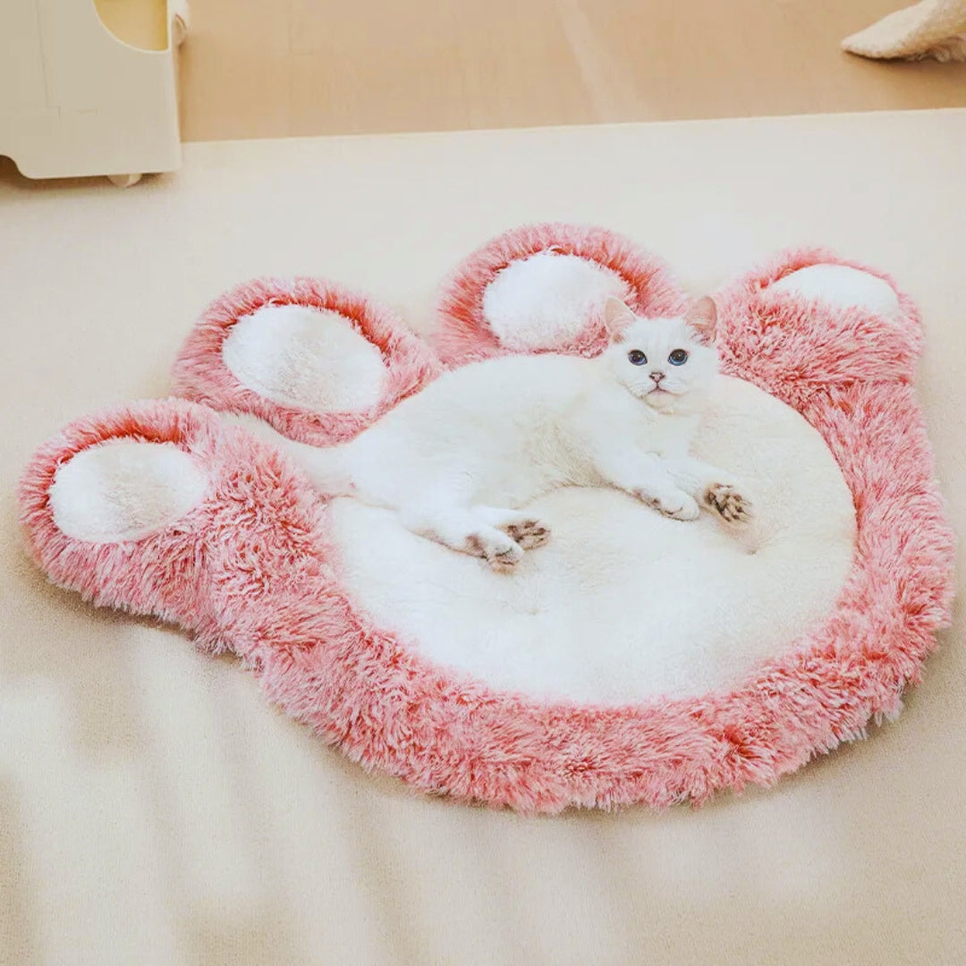 A white cat lying on the pink paw-shaped plush mat on a bed