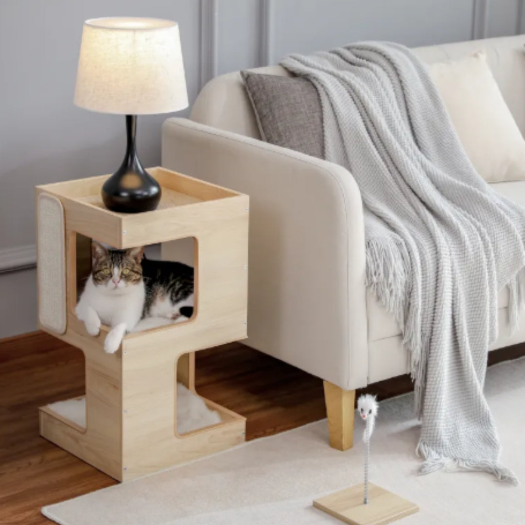 Two-tier wooden cat house beside a sofa holding a lamp, a tabby and white cat resting in the middle level
