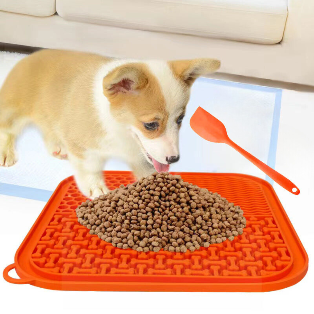 A puppy licking food from the orange silicone lick mat, the orange spatula beside it