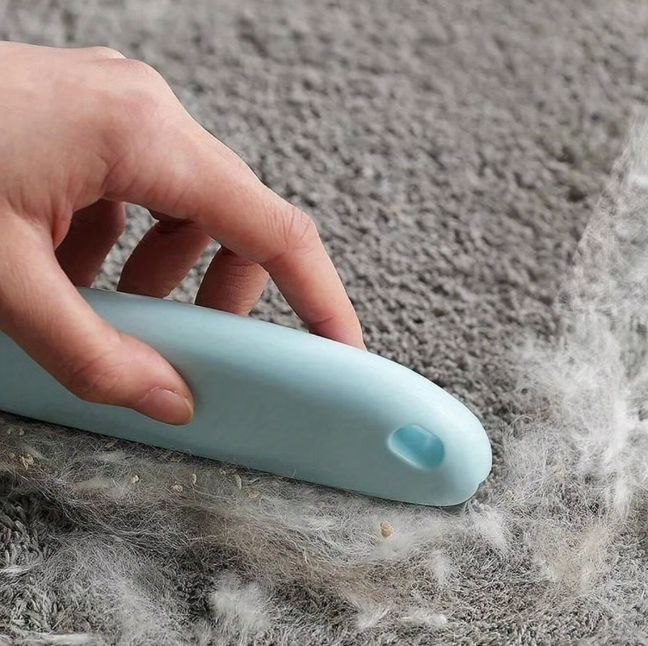 A hand drawing the light blue pet hair remover across a grey rug, gathering pale hair