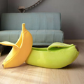 A green banana cat bed on a wooden floor beside a yellow one standing on its end