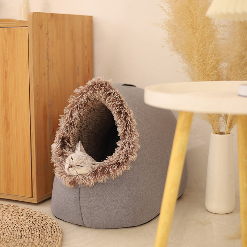 A cat resting inside the grey cave bed with a brown-tipped plush entrance, beside a wooden cabinet