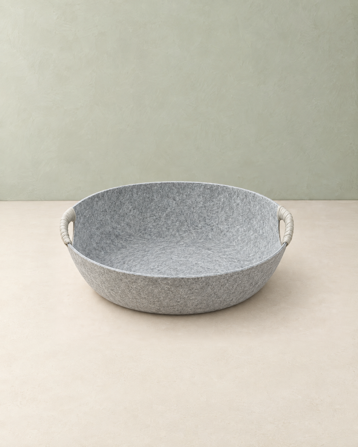 Light grey felt cat bed basket with two rope-wrapped handles on a cream floor against a pale sage wall