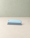 Light blue hand-held pet hair remover with a grey toothed blade, on a cream floor against a pale sage wall