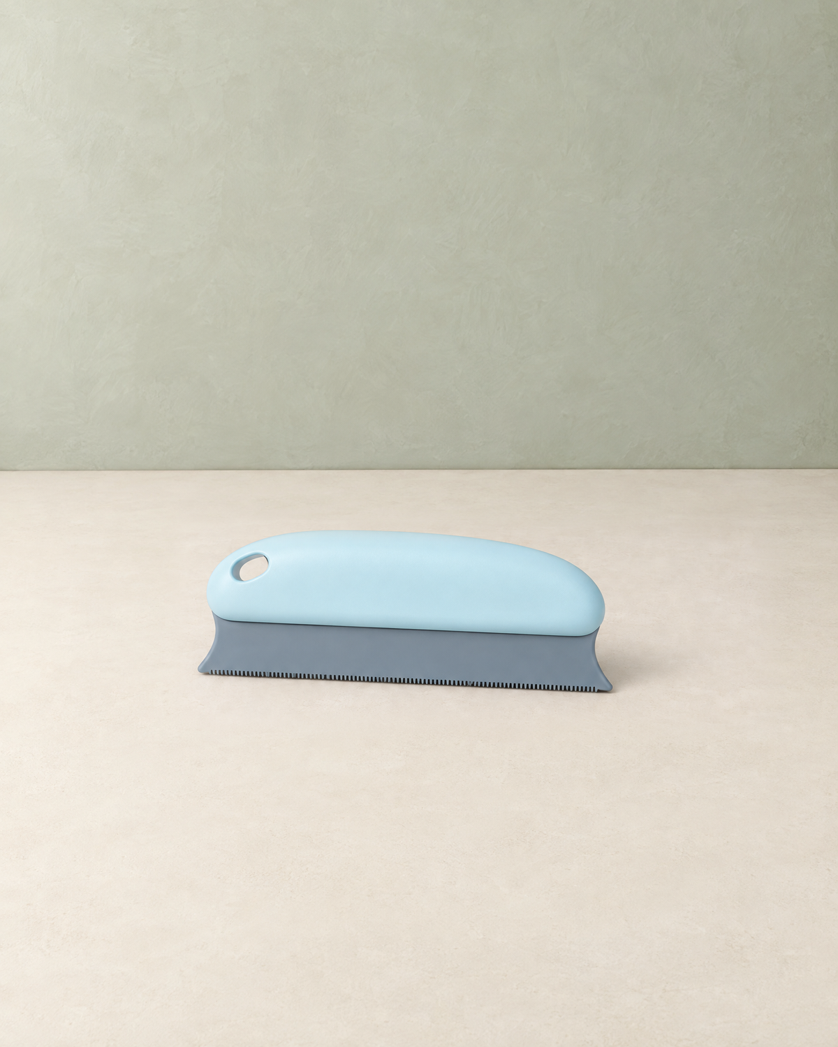 Light blue hand-held pet hair remover with a grey toothed blade, on a cream floor against a pale sage wall