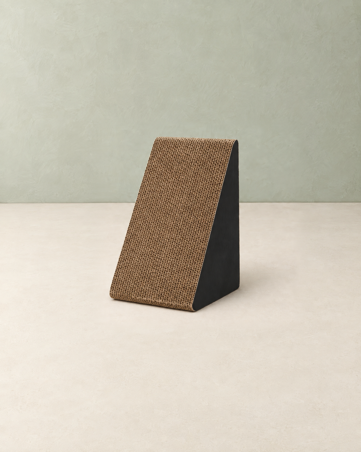 Small triangular cat scratcher with a brown corrugated cardboard face and black sides, on a cream floor against a pale sage wall