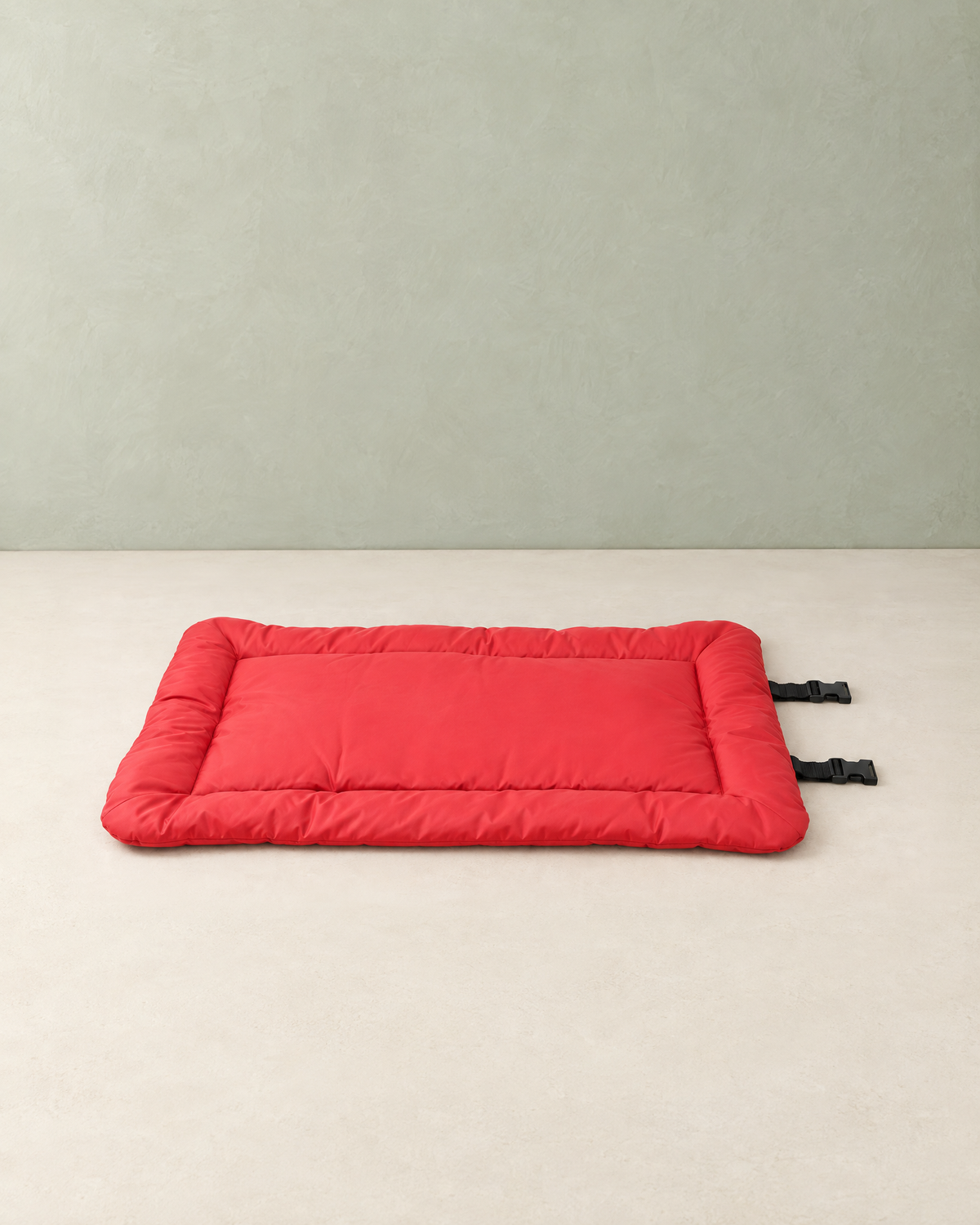 Red roll-up travel dog bed lying flat on a cream floor against a pale sage wall, two black buckle straps at one end