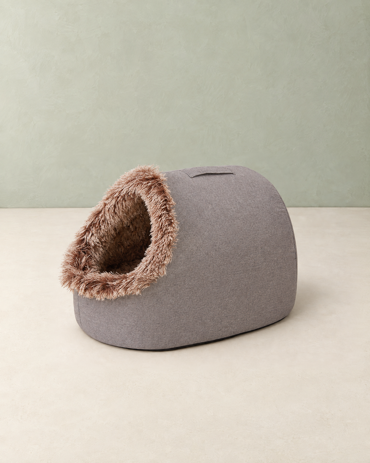 Grey cave dog bed with a fluffy brown-tipped plush entrance on a cream floor against a pale sage wall