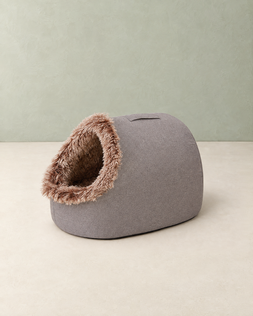 Grey cave dog bed with a fluffy brown-tipped plush entrance on a cream floor against a pale sage wall