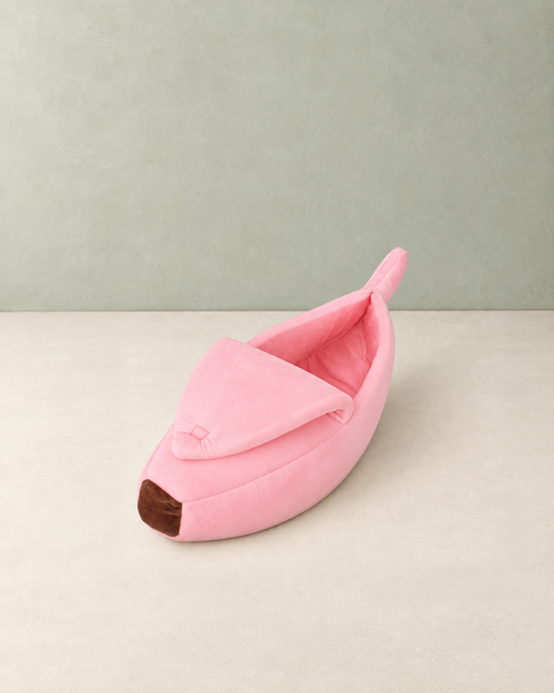Pink banana-shaped plush cat bed with a brown tip and the peel folded over the front, on a cream floor against a pale sage wall