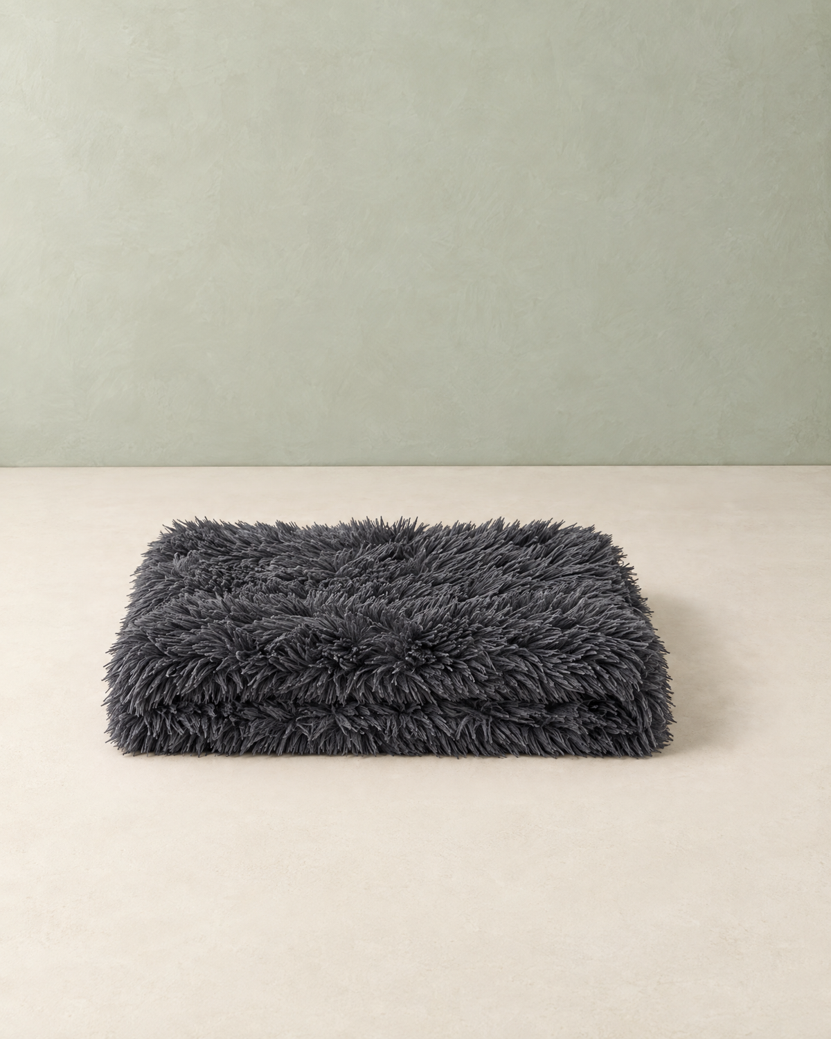 Dark grey long-pile plush dog blanket, folded, on a cream floor against a pale sage wall