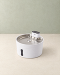 White round pet water fountain with a stainless steel tray and a control module on top, on a cream floor against a pale sage wall
