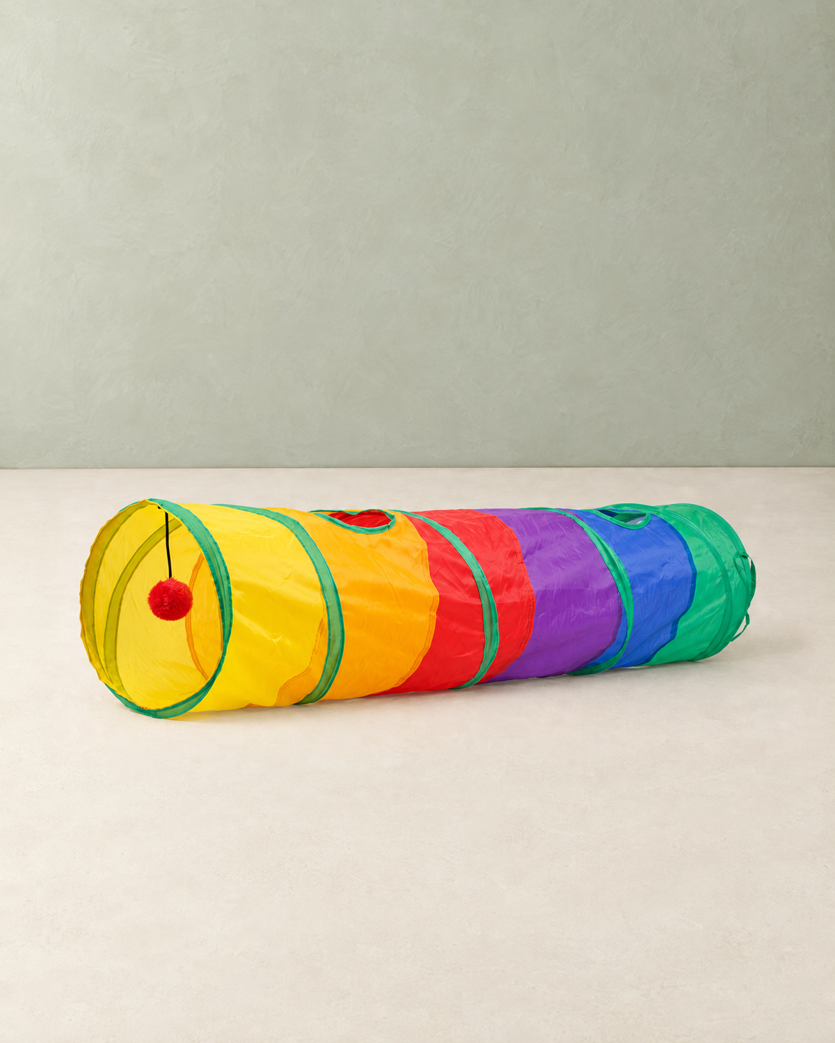 Rainbow cat tunnel lying on its side on a cream floor, a red pom-pom hanging inside the yellow entrance