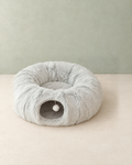 Light grey plush cat tunnel bed shaped like a ring, with a round tunnel opening and a white pompom, on a cream floor against a pale sage wall