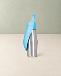 Stainless steel dog water bottle with a light blue trough folded along its side, standing on a cream floor against a pale sage wall