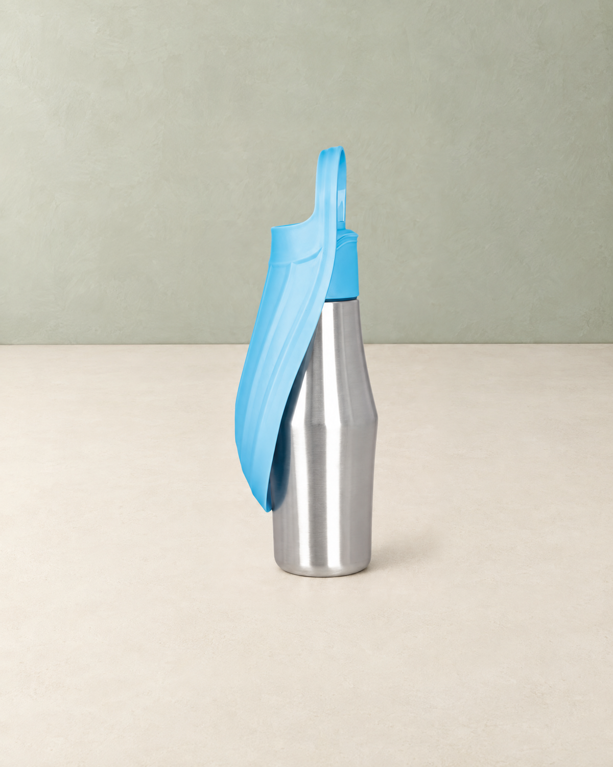 Stainless steel dog water bottle with a light blue trough folded along its side, standing on a cream floor against a pale sage wall