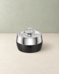 Stainless steel pet water fountain with a T-shaped double spout and a black ribbed base, on a cream floor against a pale sage wall