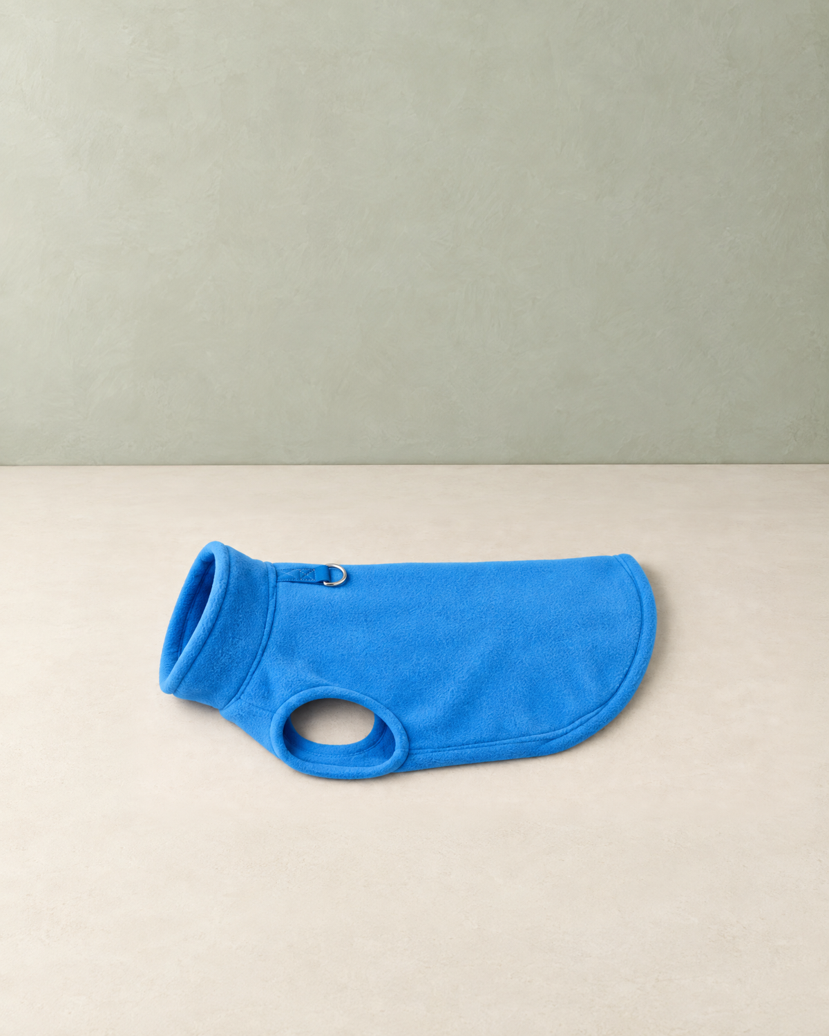 Blue polar fleece dog vest with a rolled collar and a D-ring, lying flat on a cream floor against a pale sage wall