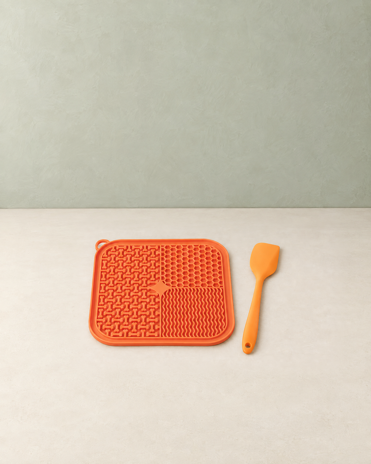 Orange silicone lick mat with its orange spatula lying on a cream floor against a pale sage wall