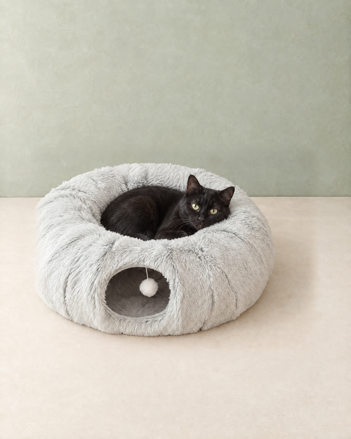 A black cat curled up in the middle of the light grey plush cat tunnel bed